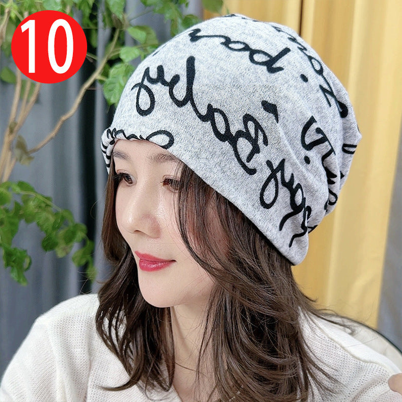 Woman wearing a gray beanie with black text, sitting outdoors with plants in the background.