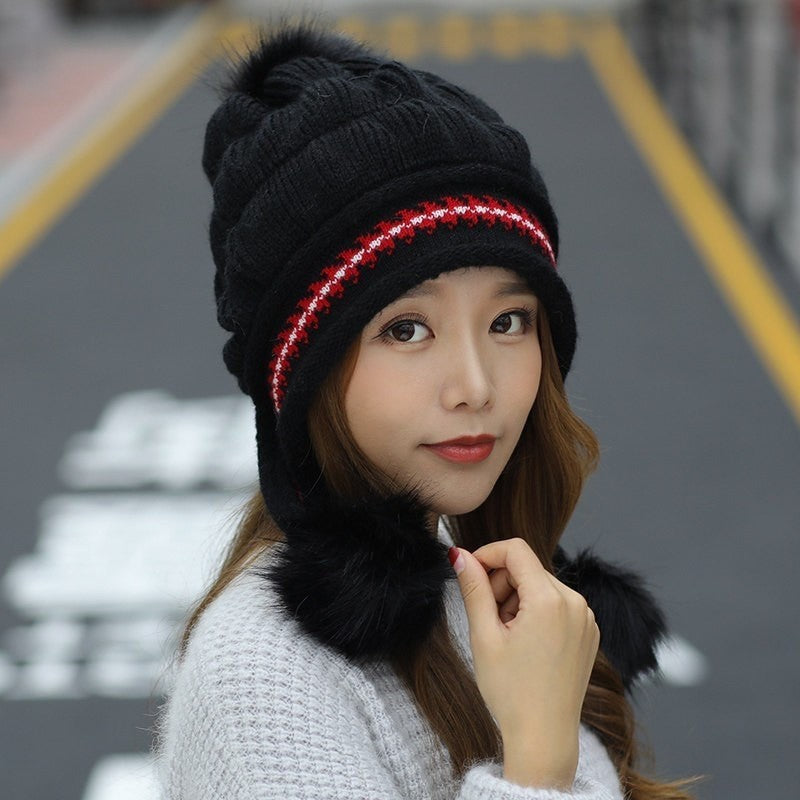Woman wearing a black knit beanie with red accents and fur trim, standing on a blurred street background.