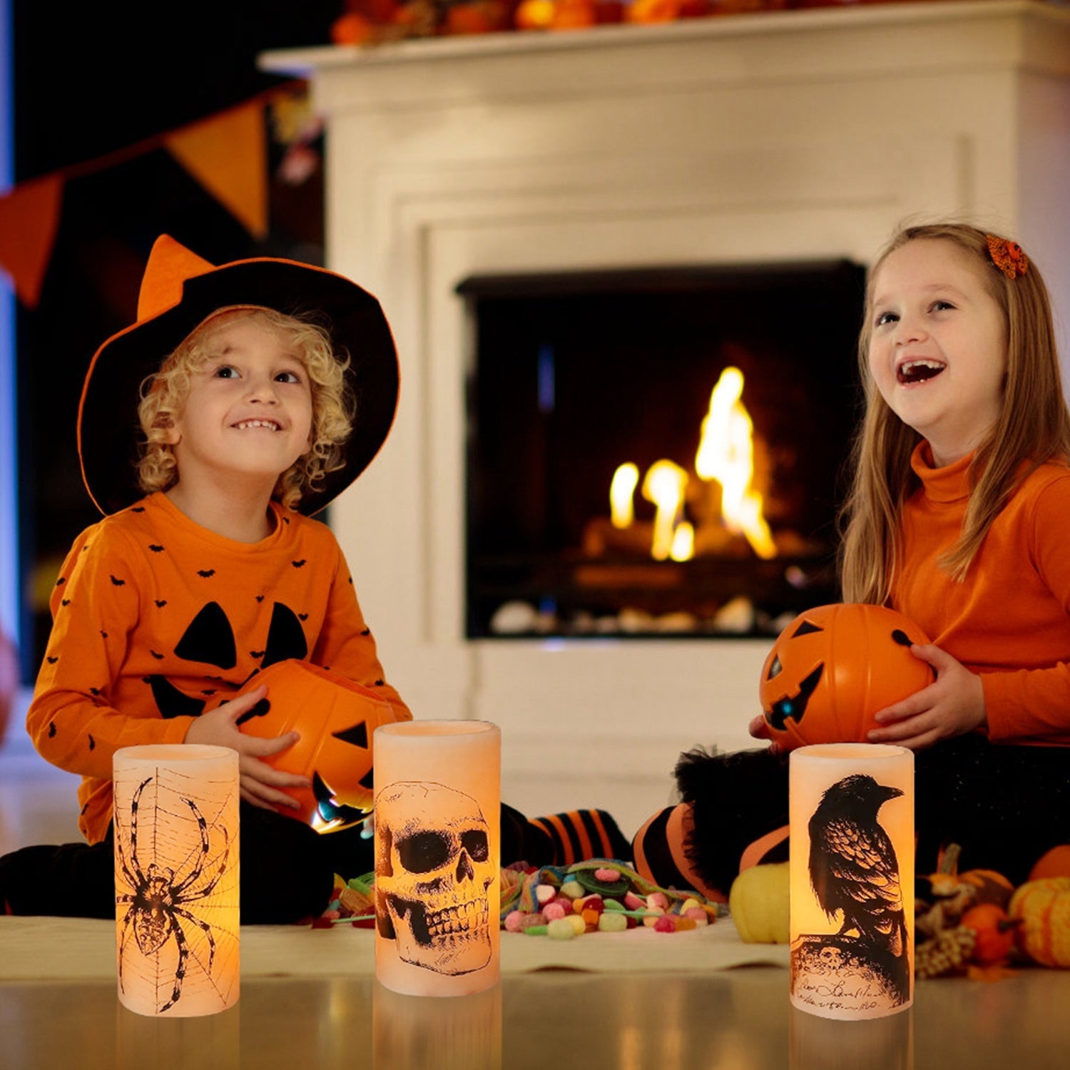 Two children in Halloween costumes sitting in front of a fireplace with lit candles and pumpkins.